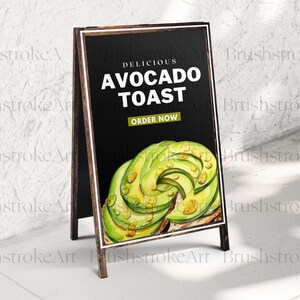 Op de afbeelding: A-frame schoolbordbord met de tekst "Delicious Avocado Toast". Het bord bevat een aquarel illustratie van avocadoschijfjes en de woorden "Order Now". Het bord heeft een donkere achtergrond en een houten frame.