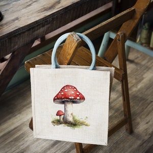 May include: A beige tote bag with a blue handle and a red and white mushroom illustration. The bag is hanging on a wooden chair.