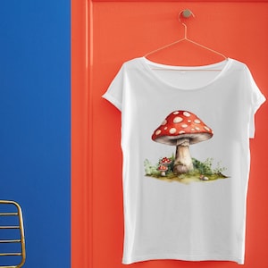May include: White t-shirt with a red and white mushroom graphic. The mushroom has white spots and is surrounded by green foliage.