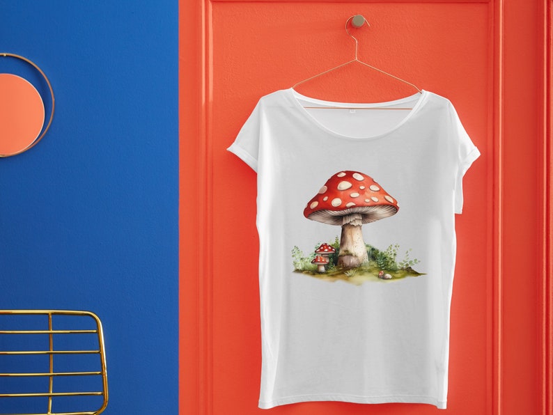 May include: White t-shirt with a red and white mushroom graphic. The mushroom has white spots and is surrounded by green foliage.