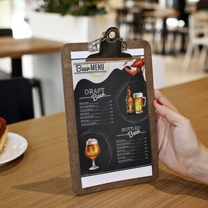 Op de afbeelding: Een bruin clipboard met een wit papieren menu met een lijst van bieren van de tap en in flessen. Het menu heeft een handgetekende illustratie van een bierglas en een fles. Het clipboard staat op een houten tafel met een stuk aardbeiencheesecake op de voorgrond.