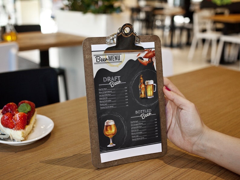 Op de afbeelding: Een bruin clipboard met een wit papieren menu met een lijst van bieren van de tap en in flessen. Het menu heeft een handgetekende illustratie van een bierglas en een fles. Het clipboard staat op een houten tafel met een stuk aardbeiencheesecake op de voorgrond.