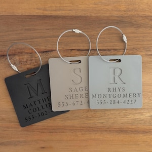 May include: Three personalized luggage tags in black, beige, and gray, each with a silver wire loop. The tags display a single initial, a name, and a phone number. The tags are square with rounded corners.