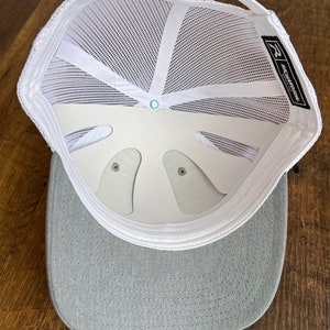 Pu&ograve; includere: Un cappello da camionista in rete bianca con visiera grigia. Il cappello ha una cinghia regolabile bianca e un'etichetta nera con il testo "Richardson 112" sul retro.