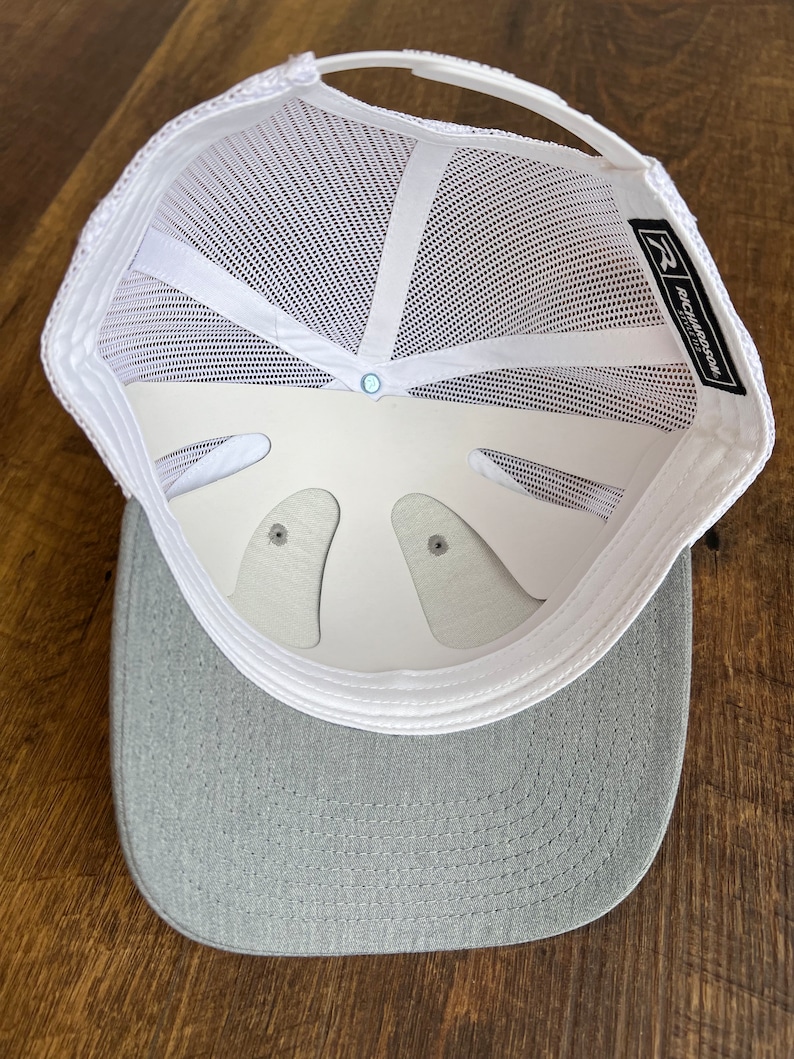 Pu&ograve; includere: Un cappello da camionista in rete bianca con visiera grigia. Il cappello ha una cinghia regolabile bianca e un'etichetta nera con il testo "Richardson 112" sul retro.