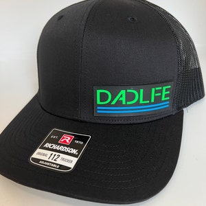 May include: Black trucker hat with a mesh back and a curved brim. The hat features the text "DADLIFE" in green and blue on a black rectangular patch. A white tag with "RICHARDSON" and "ORIGINAL 112 TRUCKER" is attached.