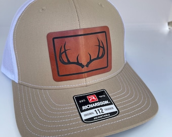 Puede incluir: Un gorro de camionero de color beige y blanco con un parche de cuero marrón que presenta una silueta de una cornamenta de ciervo. El gorro tiene una parte posterior de malla blanca y una correa ajustable negra con el texto "Richardson 112 Original Trucker Adjustable" en una etiqueta blanca.