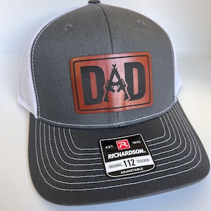 May include: A gray and white trucker hat with a brown leather patch that says "DAD" with a silhouette of two rifles crossed inside the letters. The hat has a white mesh back and a black and red label that says "Richardson" with the text "Original 112 Trucker Adjustable".