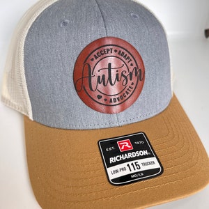 May include: A gray and white trucker hat with a brown leather patch that says "Accept Adapt Autism Advocate". The hat has a brown brim and a black label that says "EST. 1970 RICHARDSON LOW-PRO 115 TRUCKER MD/LG".