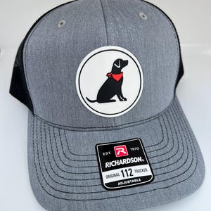 May include: Gray and black trucker hat with a white circle patch featuring a black dog wearing a red bandana. The patch also includes the text "Richardson" and "112". The hat is adjustable.