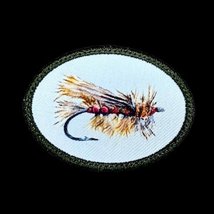 May include: Oval patch featuring a detailed embroidered fishing fly. The fly has brown and tan wings, a red body, and a black hook. The patch is bordered by a green stitched edge and set against a white background.