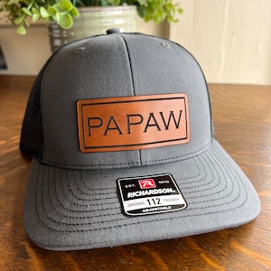 May include: A gray and black trucker hat with a brown leather patch that says "PAPAW". The hat is adjustable and has a Richardson 112 logo.