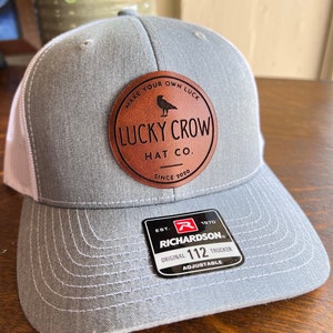 May include: Gray and white trucker hat with a brown leather patch that says "Make Your Own Luck Lucky Crow Hat Co. Since 2020". The hat also has a black and red label that says "Est. 1870 Richardson Original 112 Trucker Adjustable".