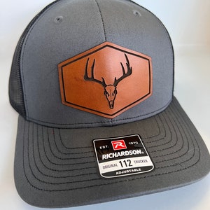 May include: A gray and black trucker hat with a brown leather patch featuring a deer skull with antlers. The patch is in the shape of a hexagon. The hat has an adjustable strap and the text "EST. 1970 RICHARDSON. ORIGINAL 112 TRUCKER ADJUSTABLE" is printed on a black label.