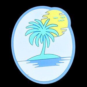 May include: A tropical island scene in a blue and white oval. A light blue palm tree sits on a small island with a yellow sun and blue water. The design is outlined in a darker blue.