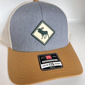 May include: A gray and brown trucker hat with a white mesh back. The hat has a patch with a moose silhouette inside a diamond shape. The patch is green and white. The hat has a black label that says "EST. 1970 RICHARDSON LOW-PRO 115 TRUCKER MD/LG".