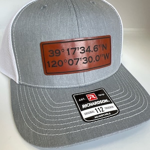 May include: A gray and white trucker hat with a brown leather patch that reads "39°17'34.6"N 120°07'30.0"W". The hat also has a black and red label that reads "EST. 1970 RICHARDSON ORIGINAL 112 TRUCKER ADJUSTABLE".