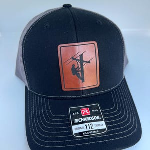 May include: Black and gray trucker hat with a brown leather patch featuring a silhouette of a lineman working on a power line. The patch has the text "Richardson" and "Est. 1970". The hat is adjustable and has the text "Original 112 Trucker" on the label.