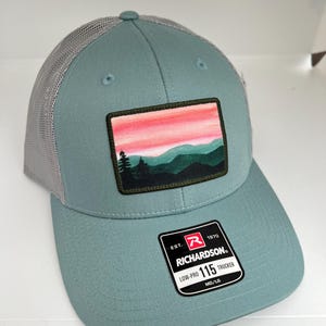May include: A light blue trucker hat with a gray mesh back. The hat has a patch on the front with a sunset scene and mountains. The hat also has a red and white label with the text "Richardson" and "115 Low-Pro Trucker".