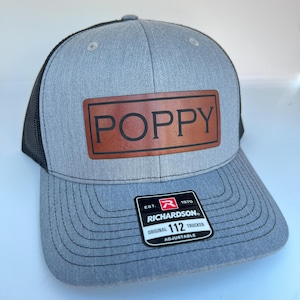 May include: Gray trucker hat with a black mesh back. The front features a brown leather patch with the name "POPPY" in capital letters. A black and white tag reads "RICHARDSON ORIGINAL 112 TRUCKER ADJUSTABLE."