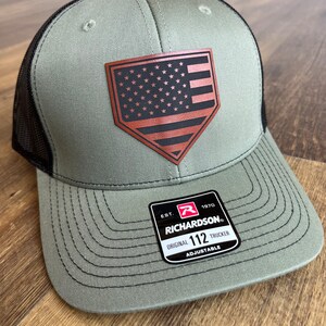 May include: A green and black baseball cap with a brown leather patch featuring an American flag. The patch is shaped like a baseball diamond. The cap has a black mesh back and an adjustable strap with the text "EST. 1970 RICHARDSON. ORIGINAL 112 TRUCKER ADJUSTABLE".