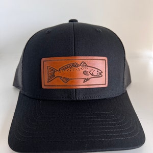 May include: A black trucker hat with a brown leather patch featuring a detailed illustration of a fish. The patch is centered on the front of the hat.