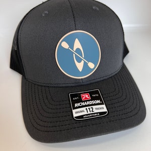May include: A dark gray trucker hat with a blue and white kayak patch on the front. The hat has a black mesh back and a brown adjustable strap with the text "EST. 1970 RICHARDSON. ORIGINAL 112 TRUCKER ADJUSTABLE".