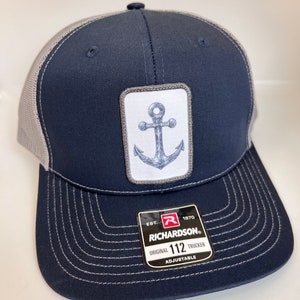 May include: A navy blue and gray trucker hat with a white anchor graphic and a black and red Richardson label. The label says "Est. 1870 Richardson Original 112 Trucker Adjustable".