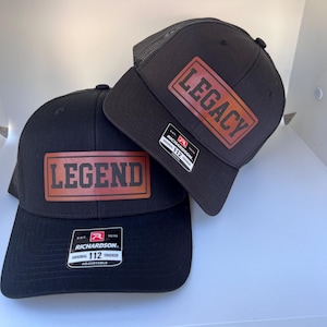 May include: Two black trucker hats with brown leather patches. The patch on the left hat says "LEGEND" and the patch on the right hat says "LEGACY".