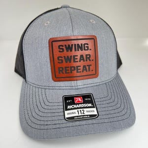 May include: A gray and black trucker hat with a brown leather patch that says "Swing.  Swear. Repeat."  The hat is adjustable and has a black mesh back.  The hat has a red and black Richardson logo with the text "EST. 1970 RICHARDSON ORIGINAL 112 TRUCKER ADJUSTABLE".