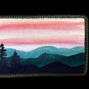 May include: Rectangular patch featuring a landscape scene. The design depicts a mountain range in shades of teal and navy, under a pink and orange sky. A dark green border frames the image, with a silhouetted tree in the foreground.