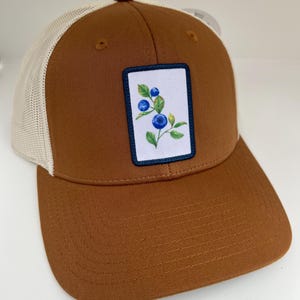 May include: A brown and white trucker hat with a patch featuring a blue and green blueberry sprig. The hat has a mesh back.