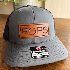 May include: A gray and black trucker hat with a brown leather patch that says "POPS" on it. The hat is adjustable and has a Richardson 112 label.