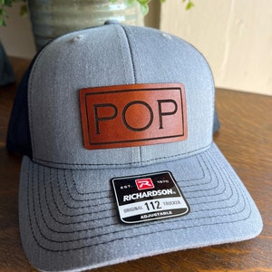 May include: Gray trucker hat with a black mesh back and a brown leather patch that reads "POP". The hat has a curved brim and an adjustable strap. The Richardson brand tag is visible.