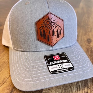 May include: Gray and white trucker hat with a brown leather patch featuring a sun, mountains, and trees. The patch is a hexagon shape. The hat has a white mesh back and a black adjustable strap with a black tag that reads "EST. 1970 RICHARDSON. ORIGINAL 112 TRUCKER ADJUSTABLE".