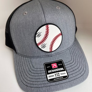 May include: Gray and black trucker hat with a red and white baseball patch. The hat has a black mesh back and an adjustable strap with the text "Richardson 112 Original Trucker Adjustable" on a black and red label.