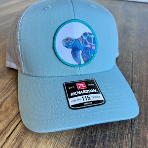 May include: A light blue trucker hat with a mesh back. The hat features a round patch with a colorful sea turtle design. The patch has a teal border. The hat has a black label with the text "EST. 1970 RICHARDSON. LOW-PRO 115 TRUCKER MD/LG".