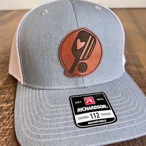 May include: Gray and white trucker hat with a brown leather patch featuring a pickleball paddle and ball. The patch has a heart on the paddle. The hat has an adjustable strap and a label that reads "Richardson. Original 112 Trucker Adjustable. Est. 1970."