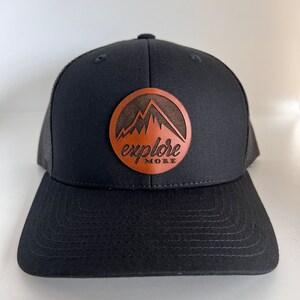 May include: Black trucker hat with a brown leather patch that says "explore more" and features a mountain silhouette.