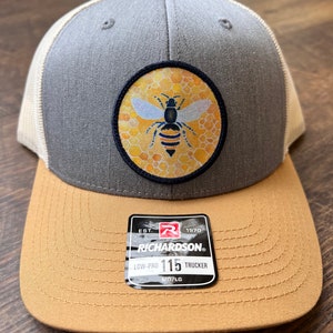 May include: A gray and tan trucker hat with a bee and honeycomb patch on the front. The hat has a black label on the side that reads "EST. 1970 RICHARDSON. LOW-PRO 115 TRUCKER MO/LG".