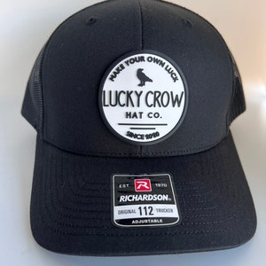 May include: Black trucker hat with a white patch that says "Make Your Own Luck Lucky Crow Hat Co. Since 2020". The patch features a black crow silhouette. The hat has a black mesh back and a black adjustable strap with a red "R" and "Richardson" logo.