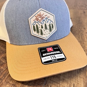 May include: A gray and tan trucker hat with a patch featuring a mountain scene with a sun and trees. The hat has a Richardson 115 Low-Pro Trucker label.
