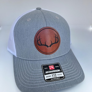 May include: Gray and white trucker hat with a brown leather patch featuring a deer antler design. The patch has the text "Richardson" and "112" on it. The hat is adjustable.