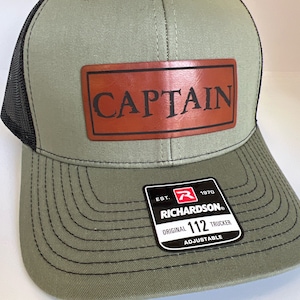 May include: A green and black trucker hat with a brown leather patch that says "CAPTAIN". The hat has a black mesh back and a Richardson 112 adjustable label.