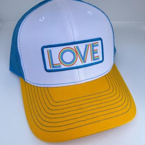 May include: A trucker hat with a white front panel, blue mesh back, and a yellow brim. The hat features the word "LOVE" in a rainbow-colored design within a blue rectangular patch.