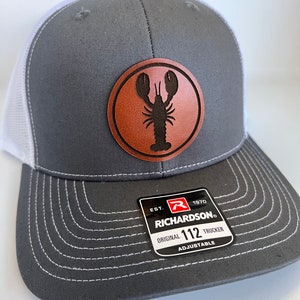 May include: Gray and white trucker hat with a brown leather patch featuring a silhouette of a lobster. The patch has the text "Richardson" and "112 Trucker" on it.
