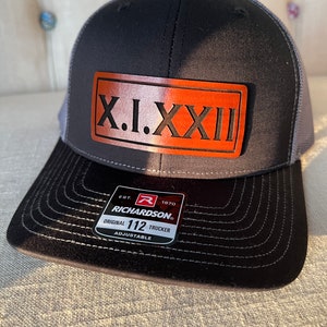 May include: A black and gray trucker hat with a brown leather patch that says "X.I.XXII". The hat has a red and white label that says "Richardson" and "Original 112 Trucker Adjustable".