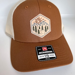 May include: A brown and white trucker hat with a patch featuring a mountain scene with a sun and trees. The hat has a white mesh back and a brown front. The hat has a label that reads "Richardson" and "Low-Pro 115 Trucker".