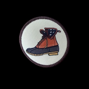 May include: Embroidered patch featuring a brown and navy blue boot with a tan sole. The boot is set against a white background with a brown border. The boot's interior has a blue and white pattern.