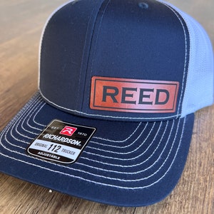 May include: A navy blue and white trucker hat with a brown leather patch that says "REED". The hat has a white mesh back and a black adjustable strap with a Richardson 112 logo.